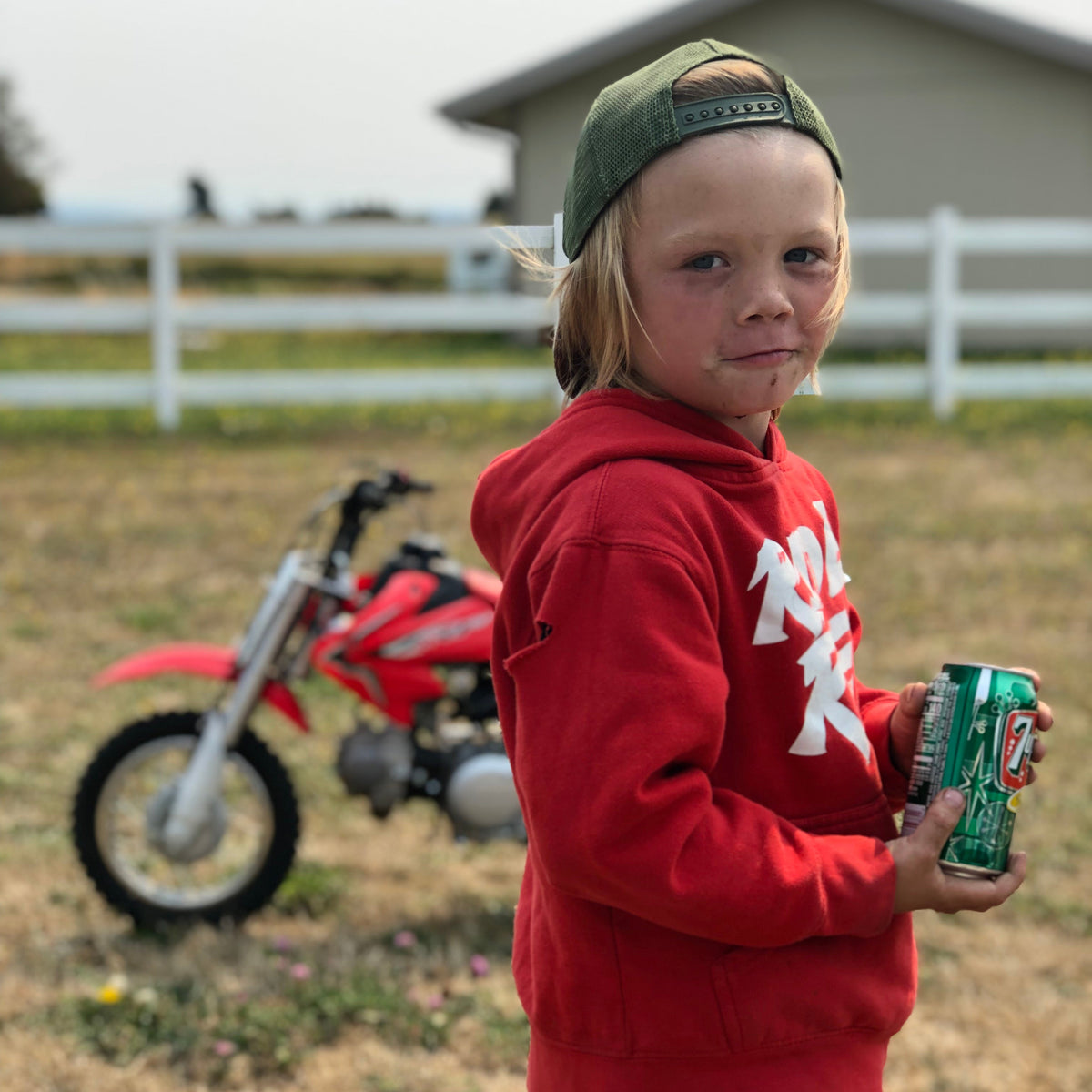 Child in a red hoodie holding a can with a dirt bike in the background