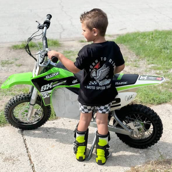 Child standing next to a green and white dirt bike on a sidewalk.