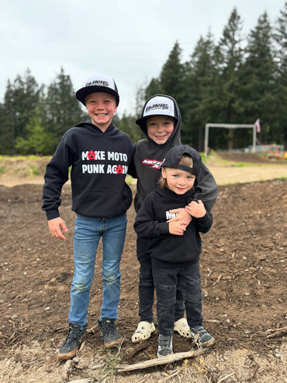 Three children in motocross gear standing on a dirt track with trees in the background.