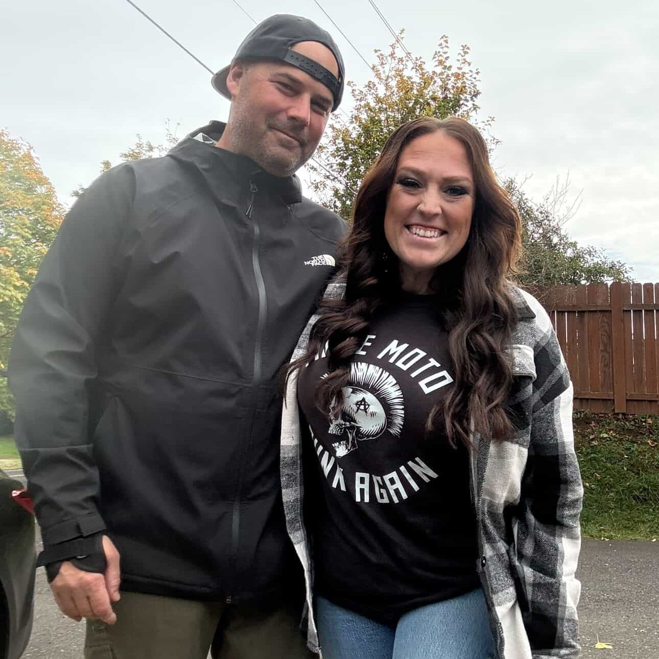 Two people standing outdoors, one wearing a black jacket and cap, the other in a black graphic t-shirt and plaid jacket.
